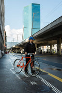 A man wearing a blue cap and black jacket stands on a city street next to a bright orange bicycle, with a tall blue glass skyscraper and an elevated highway in the background.
