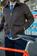 A man wearing a dark brown zip-up jacket over a light blue dress shirt and blue patterned tie, standing next to an orange bicycle in an outdoor urban setting.